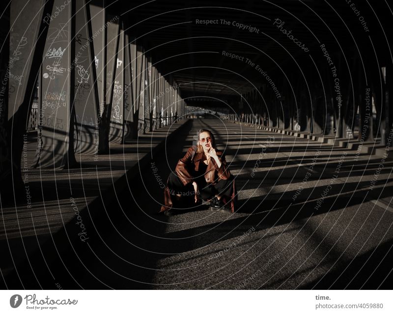 Lara Frau blick blond cool Haare lässig hand mantel ledermantel metall brücke brückenpfeiler sonnig schatten urban geheimnisvoll skeptisch straße hocken sitzen