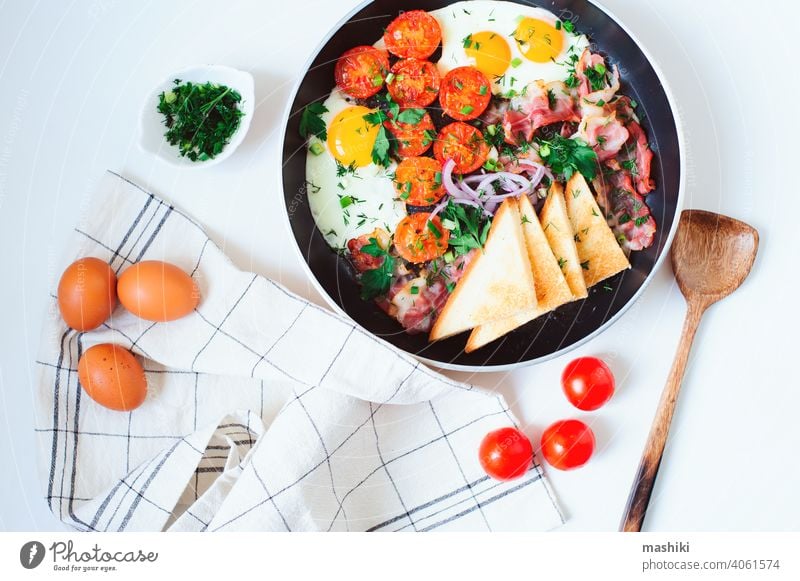 leckeres Komfort-Frühstück in der Pfanne serviert - Spiegeleier, Kirschtomaten, Speck, rote Zwiebeln und getoastetes Brot. Ei Tomate Lebensmittel Mahlzeit