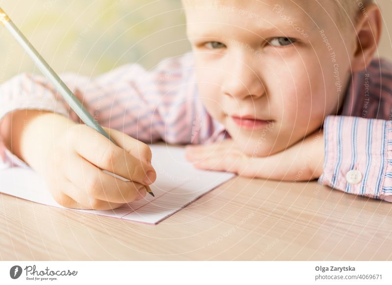 Süßer Junge schreibt seine Hausaufgaben und schaut in die Kamera. Kind lernen schreibend Tun niedlich konzentriert blond rosa Hemd Bleistift Schönschreibheft