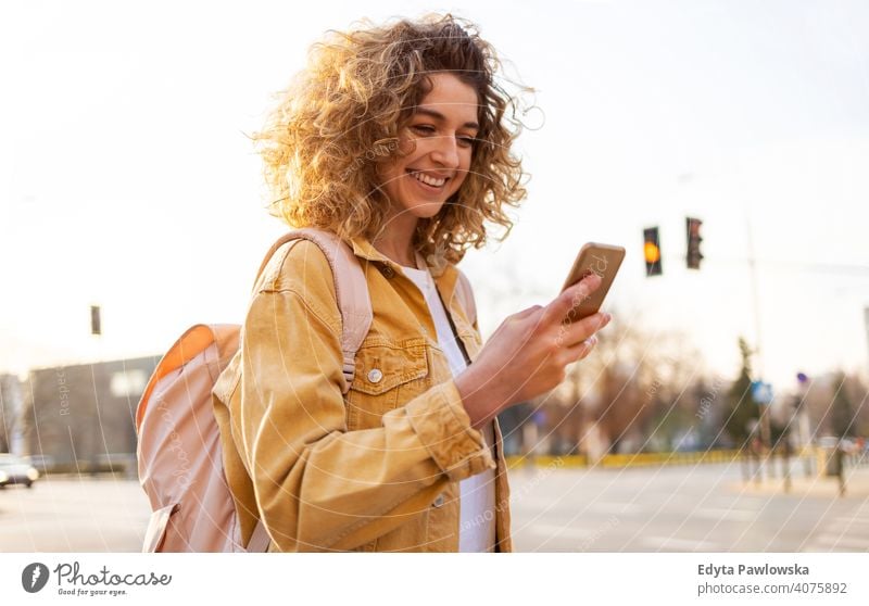 Schöne junge Frau mit lockigem Haar lächelnd und mit Smartphone natürlich Sonnenlicht urban Großstadt Hipster stylisch positiv sonnig cool Afro-Look Freude