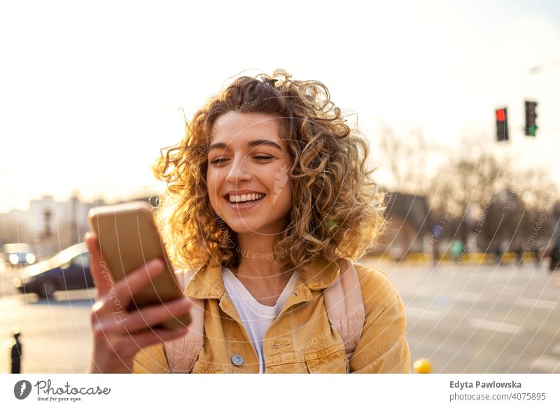 Schöne junge Frau mit lockigem Haar lächelnd und mit Smartphone natürlich Sonnenlicht urban Großstadt Hipster stylisch positiv sonnig cool Afro-Look Freude