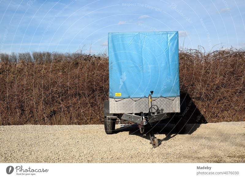 PKW Anhänger mit hoher blauer Plane aus Kunststoff vor spröder Hecke und blauem Himmel auf Kies in Naturfarben in Lemgo bei Detmold in Ostwestfalen-Lippe