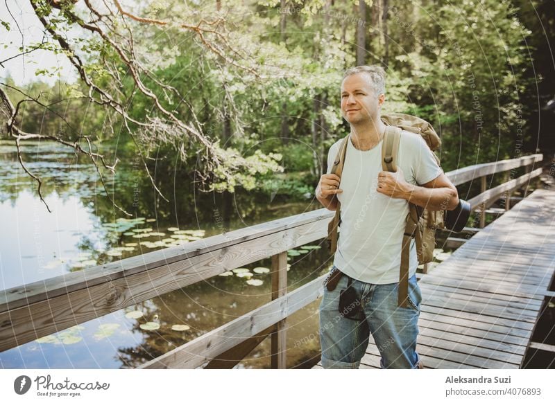 Reifer Mann erkundet die finnische Natur im Sommer, geht über die Brücke. Wanderer mit großen Rucksack Reisen in Wäldern. Sommer skandinavischen Landschaft von Seen und Wäldern.