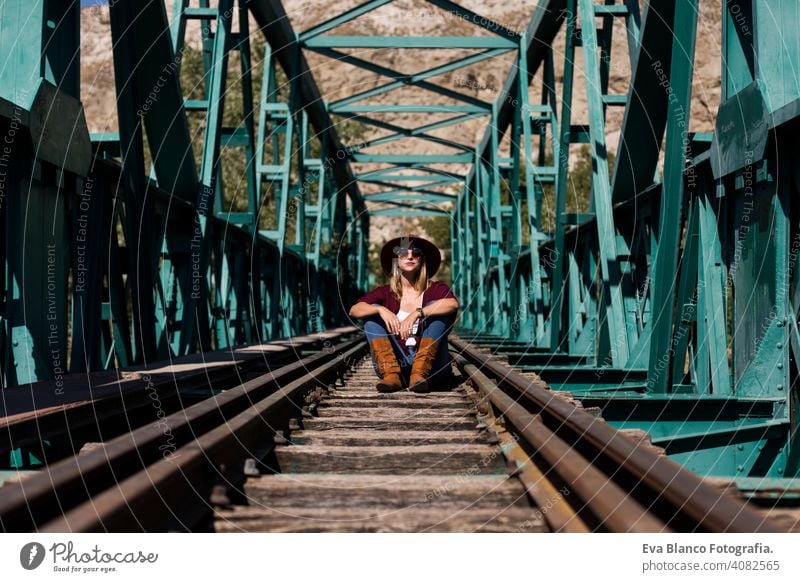 Porträt einer jungen schönen Frau, die auf der Eisenbahn einer grünen Brücke sitzt. Sie trägt stilvolle Kleidung und einen Hut. LIfestyle. Im Freien. Sonnig Zug