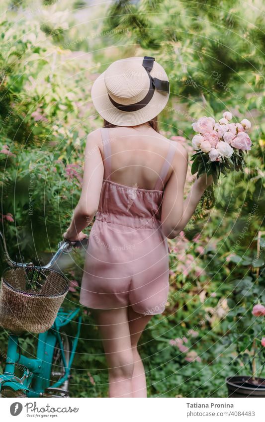 Stilvolles Mädchen in einem Strohhut und einem blassrosa pudrigen Jumpsuit geht mit einem mintfarbenen Fahrrad im Garten eines Landhauses spazieren. Sie hält einen Strauß frischer rosa Pfingstrosen in den Händen. Steht mit dem Rücken zur Kamera