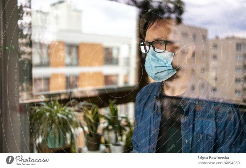 Mann mit Gesichtsmaske schaut aus dem Fenster heimwärts Haus flach Sperrung zu Hause bleiben männlich jung Erwachsener Menschen eine Person Appartement