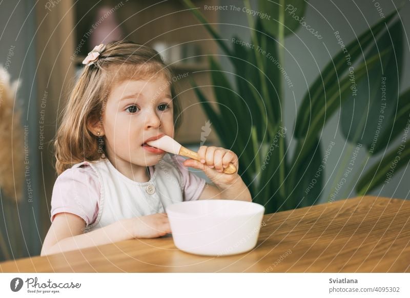 Ein kleines Mädchen isst Brei aus einer weißen Schüssel, ein Mädchen frühstückt am Tisch sitzend. Gesundes Frühstück, gesundes Essen Gesundheit Kind Haferbrei