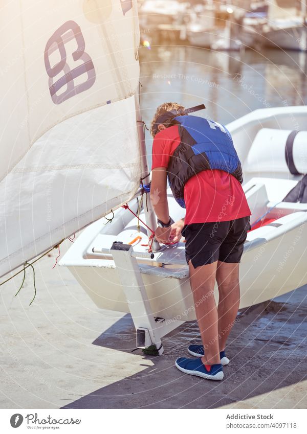 Kräftiger Junge in blauer Schwimmweste, der ein Seil verdreht und ein Segel zieht Boot Verankerung Kopplungsbindung Wasser Jacht Sicherheit Ufer Wellen Meer