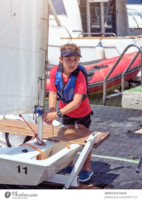 Junge in blauer Schwimmweste bindet Seil an Segelstock Kopplungsbindung Wasser Jacht Sicherheit Ufer Wellen Meer Badebekleidung Kind gespannt Training