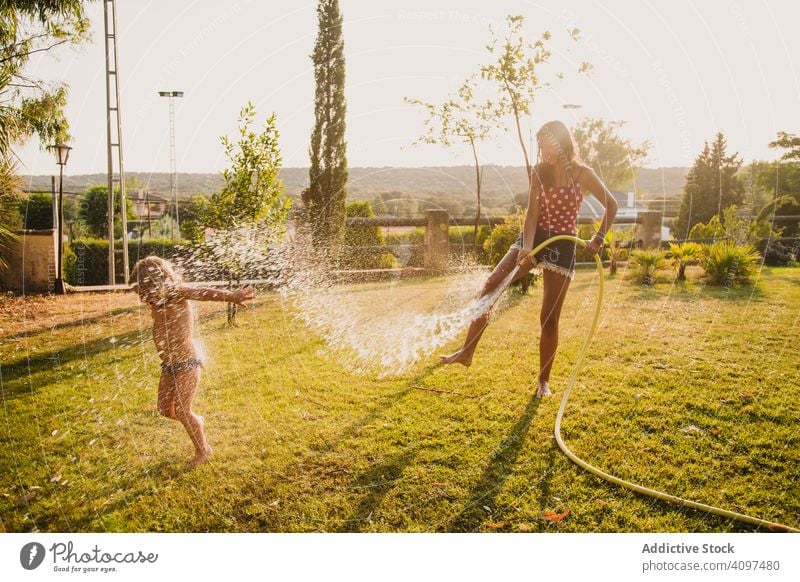 Teenager-Mädchen besprüht kleine Schwester im Garten Spaß Wasser Rasen Spray Schlauch sonnig tagsüber Hof Zusammensein Kind Glück heiter spielen Urlaub