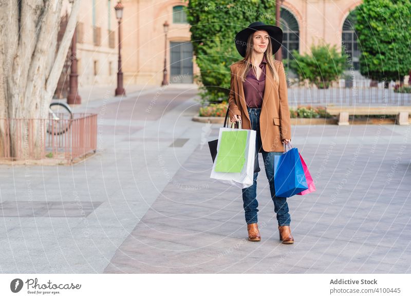 Zufriedene Frau in trendiger Kleidung beim Einkaufen in der Stadtstraße Einkaufstaschen Straße modisch schlendern Kauf Wochenende laufen stylisch Outfit Hut