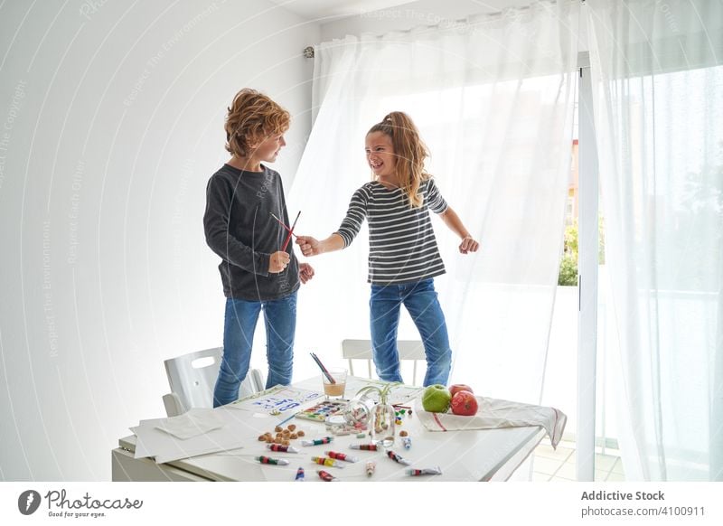 Glückliche Kinder kämpfen mit Pinseln Spaß spielen Tisch Wasserfarbe Pinselblume Freund heimwärts Kunst kreativ Zusammensein Stuhl Mädchen Farbe Junge Freude