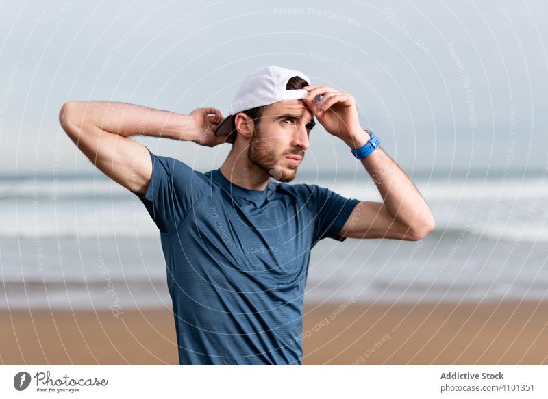 Selbstbewusster männlicher Turner in aktiver Kleidung und Mütze am Strand Sportler Training Sportkleidung Verschlussdeckel Seeküste Meeresufer Lifestyle