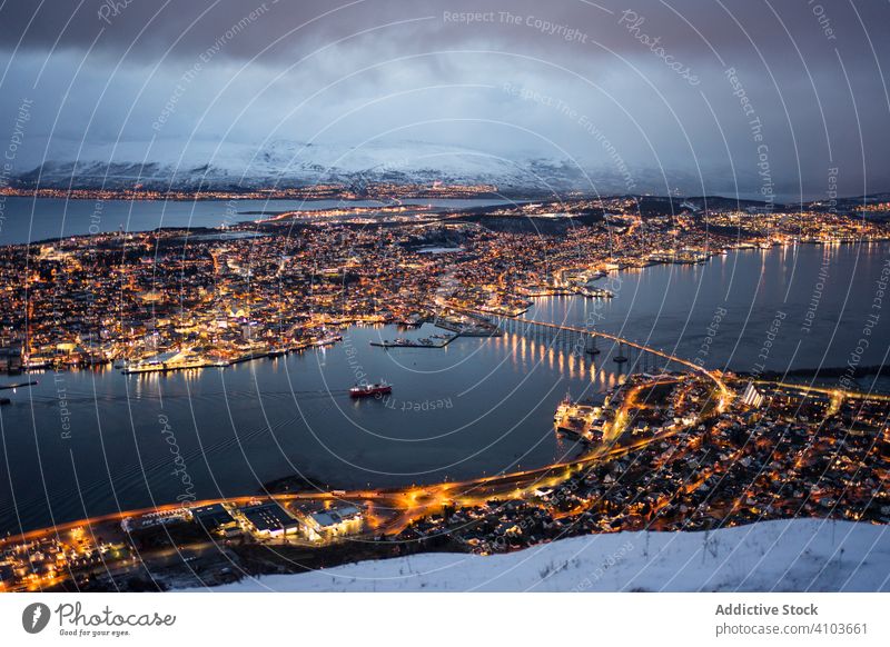 Majestätischer Blick auf die nächtliche Stadt vor verschneiten Bergen bei bedecktem Himmel Großstadt Nacht Licht Küste Hafengebiet Brücke Stadtbild golden