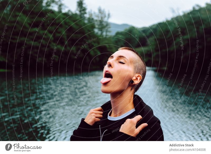 Zeitgenössische Frau in Freizeitkleidung, die sich am Regen am Teich erfreut Natur genießen fangen Regentropfen Kurze Haare Piercing lässig Skinhead informell