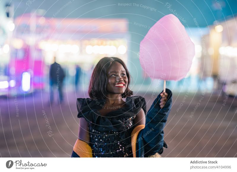 Fröhliche afroamerikanische Frau mit Zuckerwatte Messegelände Lächeln Entertainment Nacht stylisch Wochenende ethnisch jung Jahrmarkt süß Leckerbissen Dessert