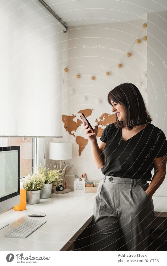 Fokussierte Frau mit dunklen Haaren, die Nachrichten über Mobiltelefone verschickt, während sie sich in der Nähe eines Desktop-Computers im Büro in Paris aufhält