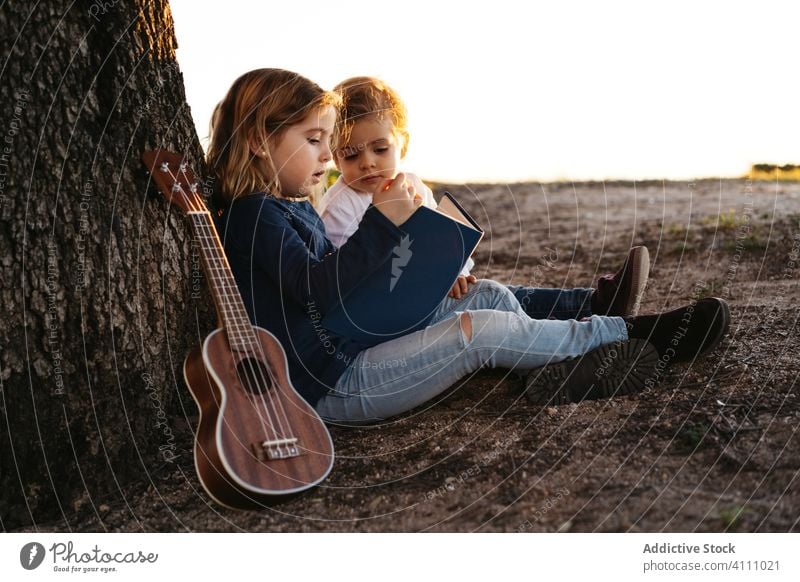 Kinder sitzen unter einem Baum und lesen ein Buch Natur Zusammensein Sommer Ukulele Gitarre ruhen ruhig Schwester Bruder Geschwisterkind Freund Landschaft