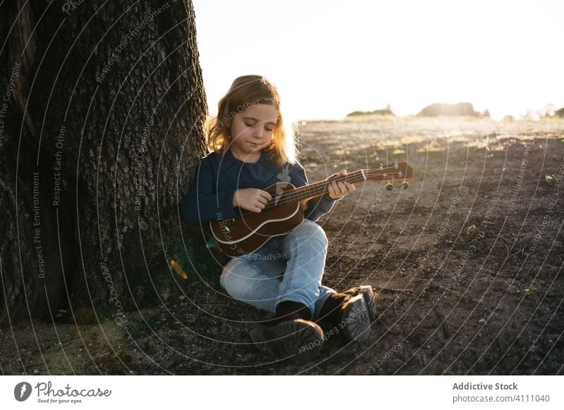 Kleines Mädchen sitzt unter einem Baum und spielt Ukulele Kind spielen Musik Gitarre Natur Sommer ernst wenig Instrument Klang Gesang Melodie sitzen Kofferraum