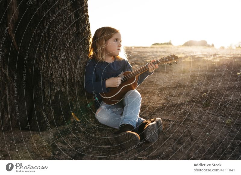 Kleines Mädchen sitzt unter einem Baum und spielt Ukulele Kind spielen Musik Gitarre Natur Sommer ernst wenig Instrument Klang Gesang Melodie sitzen Kofferraum