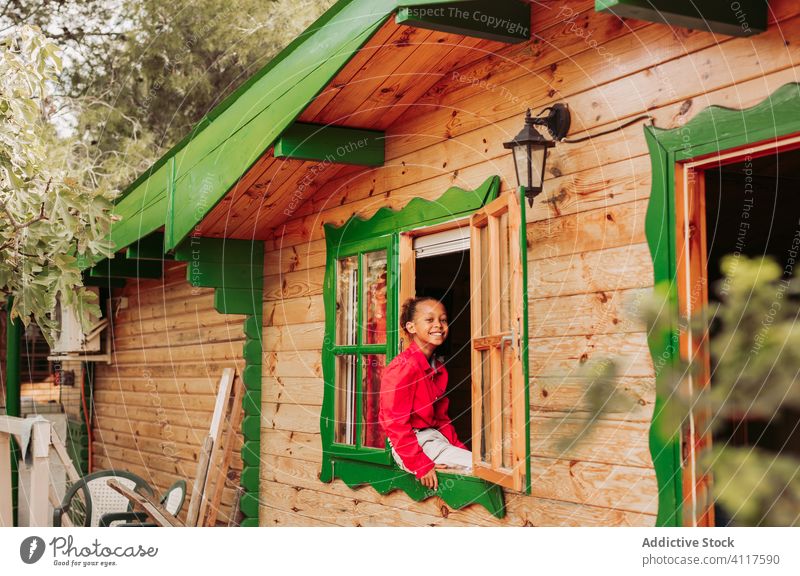 Glückliches ethnisches Kind schaut aus dem Fenster Haus heiter hölzern ländlich Lächeln heimwärts Lifestyle Landschaft Freude wenig lässig Kindheit schwarz