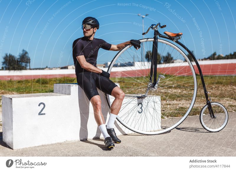 Nachdenklicher erwachsener Sportler sitzt allein auf dem Siegerpodest und stützt sich auf ein Hochrad im Sportstadion Fahrrad Podium Stadion Aspiration