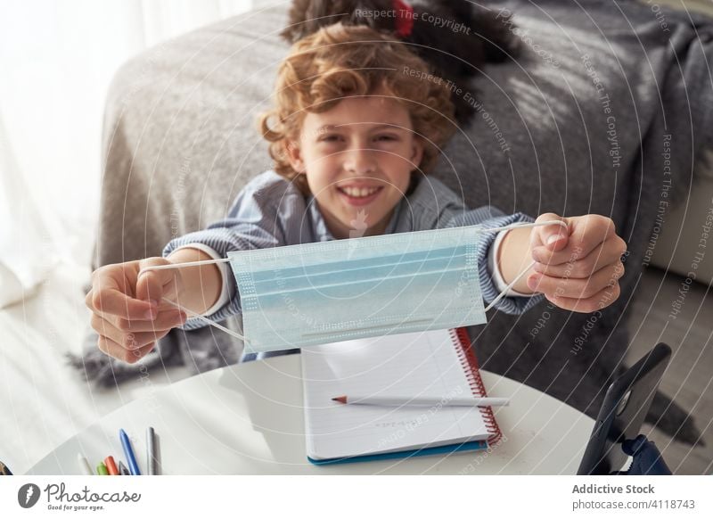 Fröhlicher Junge zeigt medizinische Maske während Fernstudium abgelegen Bildung Quarantäne Mundschutz zeigen Lächeln Notebook Smartphone heimwärts Kind lernen