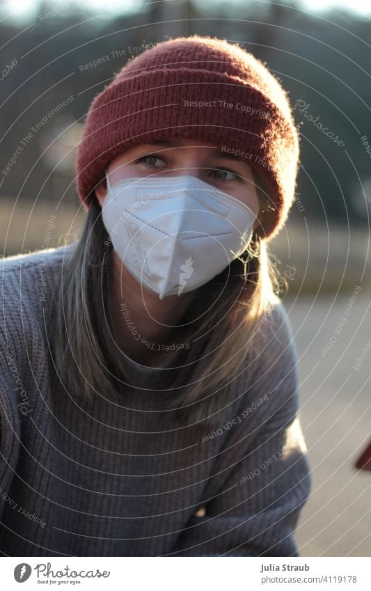 Junge Frau mit Mütze und FFP2  Atemschutzmaske sitz im Abendlicht Maske Maskenpflicht ffp2-maske ffp2 Schutzmaske coronakrise Corona-Pandemie Strickpullover