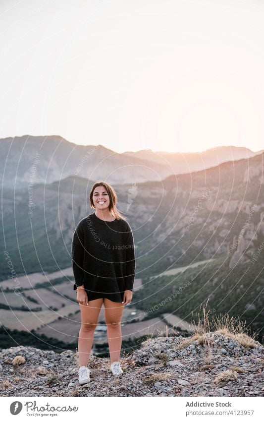 Positiver junger weiblicher Reisender, der bei Sonnenuntergang auf einem Felsen in den Bergen steht Frau Klippe Berge u. Gebirge Lachen Lächeln Stein Natur