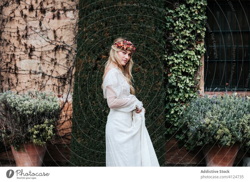 Junge Braut steht neben altem Anwesen Frau Haus Hof Buchse Wein Flora elegant Hochzeit jung Kleid Totenkranz gealtert eingetopft Blume Stil feiern Gelassenheit