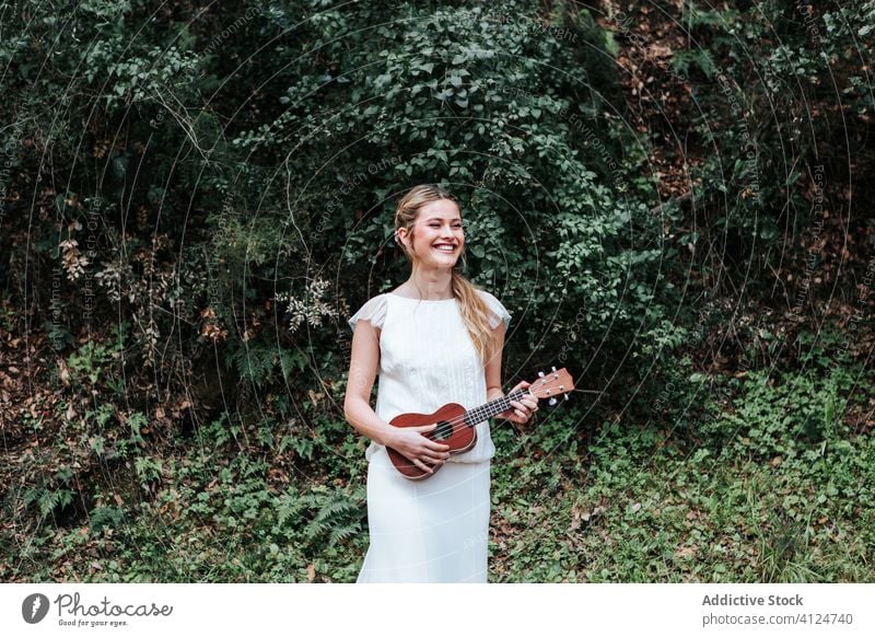 Fröhliche Braut spielt Ukulele in der Nähe von Büschen Frau spielen Landschaft Lächeln Buchse Hochzeit jung Musik Glück Kleid grün heiter Freude Sommer Natur