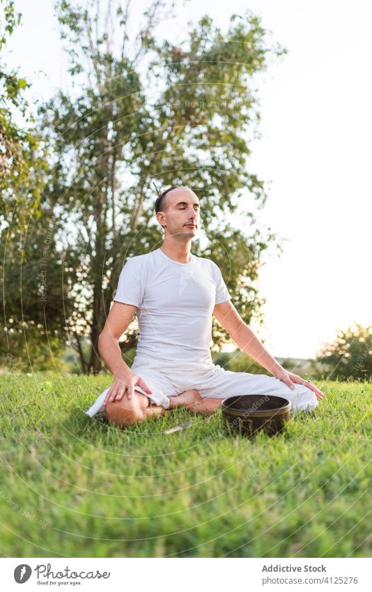 Konzentrierter junger Mann mit tibetischer Schale auf grüner Wiese Yoga tibetische Schale meditieren Klangschale Asana Windstille Zen Rasen Pose Sommer Knie