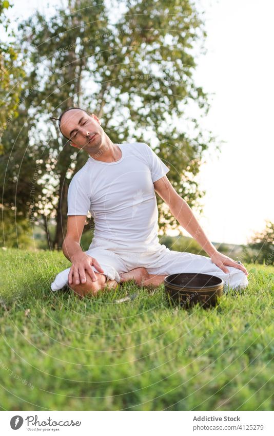 Konzentrierter junger Mann mit tibetischer Schale auf grüner Wiese Yoga tibetische Schale meditieren Klangschale Asana Windstille Zen Rasen Pose Sommer Knie