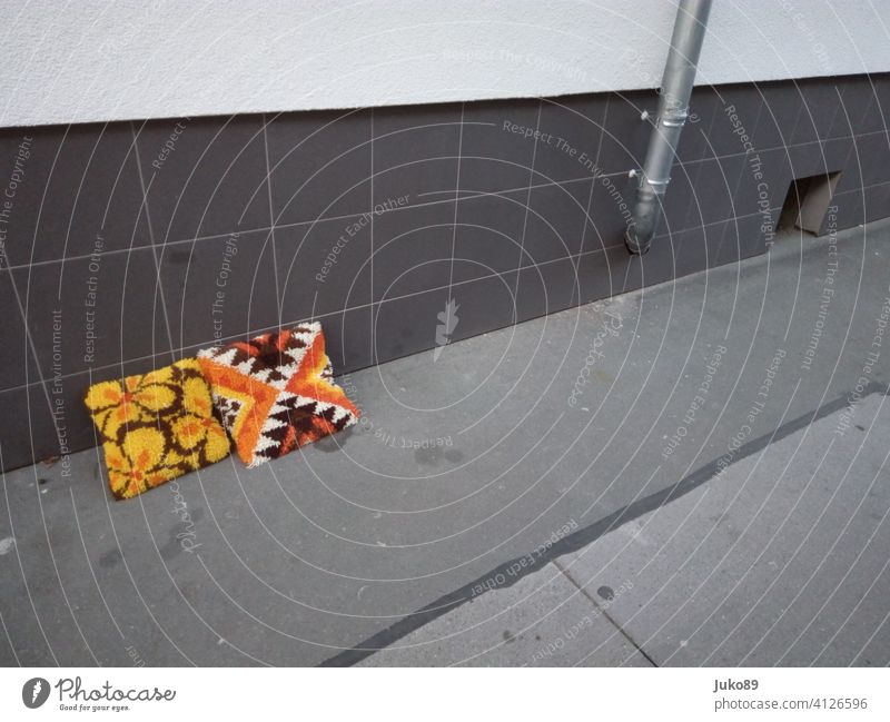 Zwei Häkelkissen im 70er-Jahre-Stil lehnen an einer grauen Hauswand auf einer Straße Kissen Kissenbezug Orange-gelb-braun retro graue Straße Regenrinne