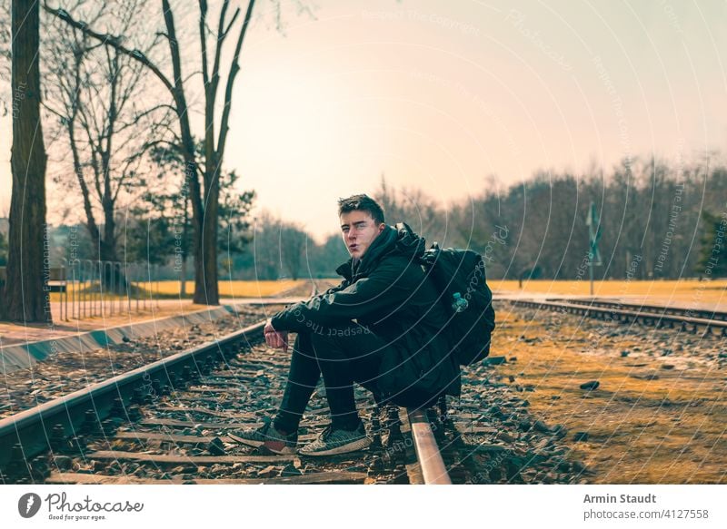 glücklicher junger Mann mit Rucksack auf Bahngleisen sitzend Sitzen Eisenbahn Spuren Landschaft im Freien Lächeln selbstbewusst kampfstark weit weg Wald Natur