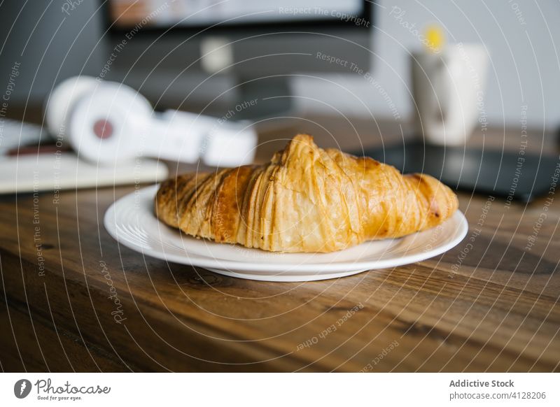 Croissant, Notizbuch und Kopfhörer auf dem Arbeitstisch Arbeitsplatz Tisch Tagebuch Monitor Morgen Frühstück Telearbeit Plan Planer Tasse Teller hölzern lecker