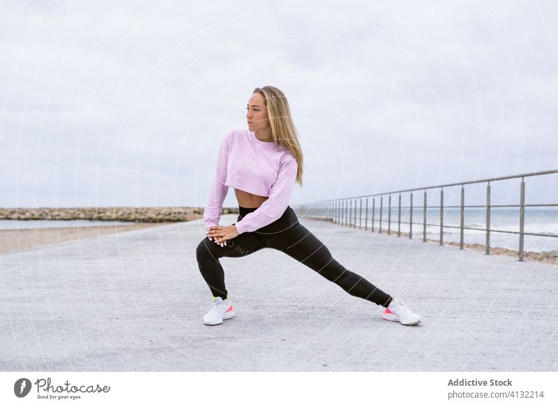 Fitnesssportlerin beim Ausfallschritt an der Böschung nahe des Ozeans Sportlerin Dehnung Bein Training Sportkleidung Stauanlage Meer Zaun Seite beweglich