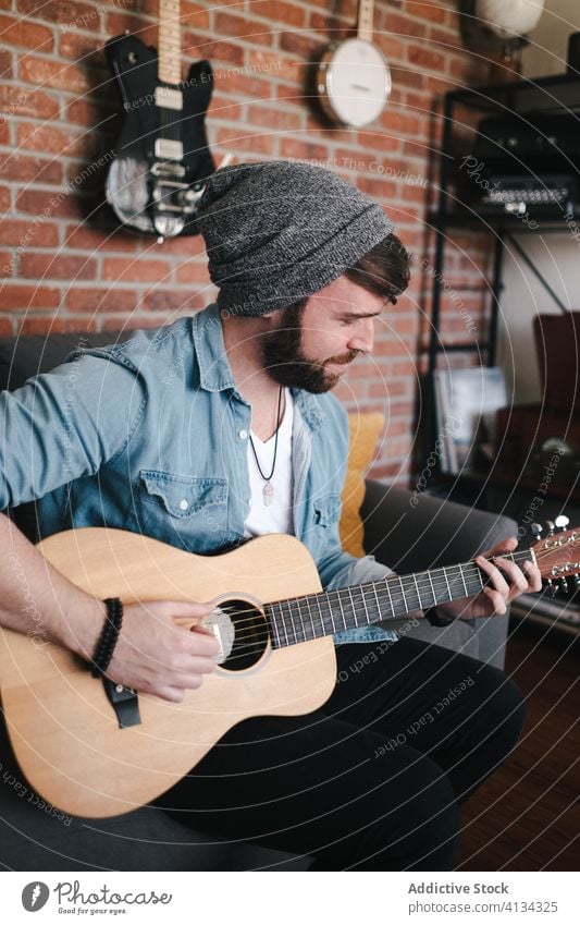 Lächelnder Musiker, der auf dem Sofa im Wohnzimmer sitzt und Gitarre spielt spielen banjo Loft akustisch Hipster Optimist flach Musical Instrument Raum bequem