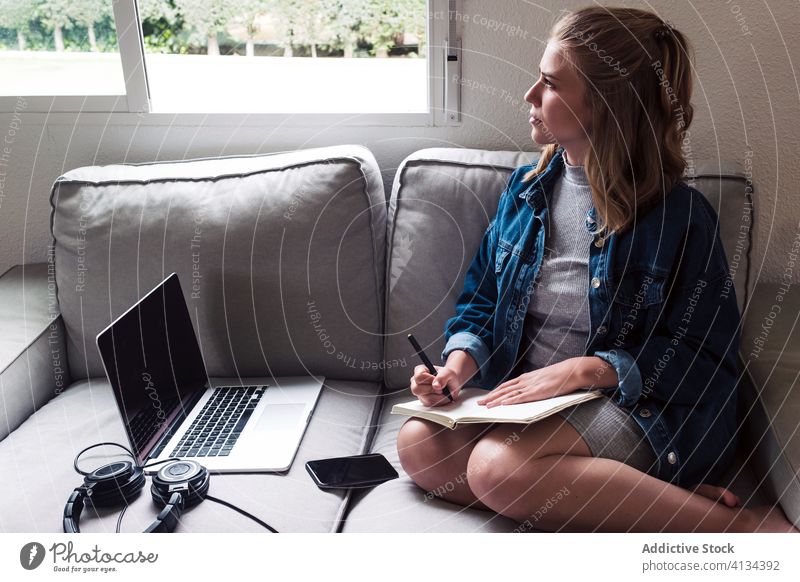 Millennial-Frau sitzt auf dem Sofa mit Gadgets und schreibt in ein Notizbuch schreiben Notebook heimwärts Apparatur jung zur Kenntnis nehmen Tagebuch