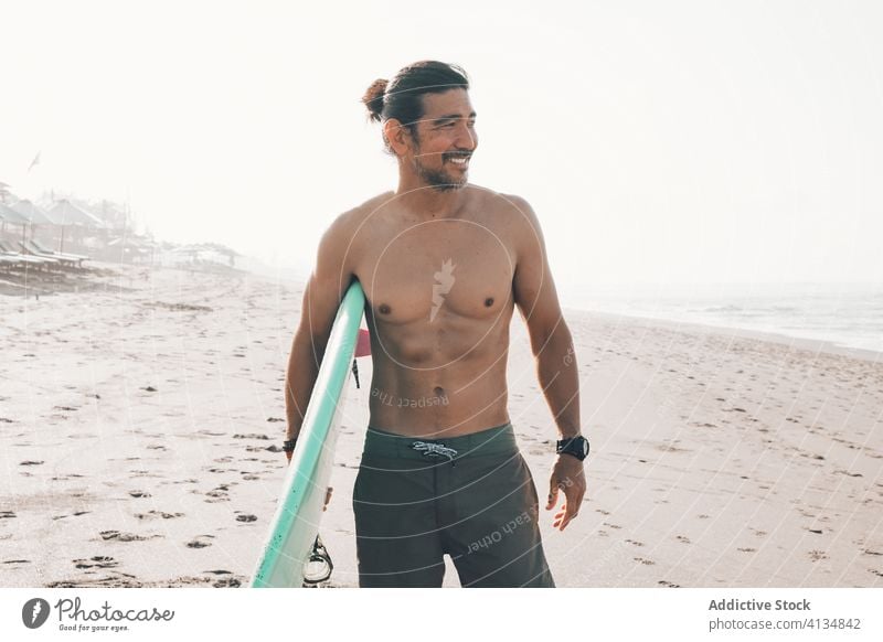 Fröhlicher Surfer mit Surfbrett am Strand stehend Mann MEER Glück aktiv Meer Küste Lächeln ohne Hemd ethnisch männlich Lifestyle Sand Sommer Ufer Urlaub Wasser