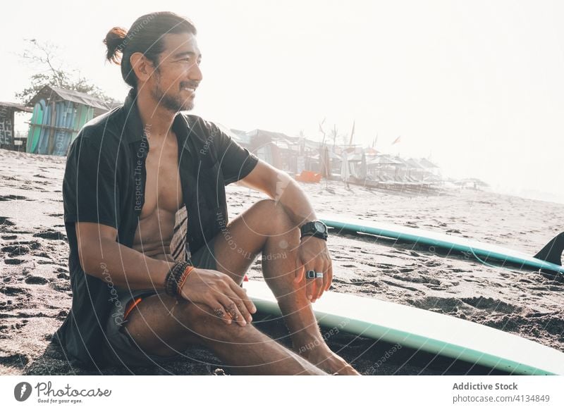 Glücklicher ethnischer Mann mit Surfbrett am Strand sitzend positiv ruhen MEER genießen Sand Sommer jung hispanisch männlich Lifestyle Urlaub Küste Meer Ufer