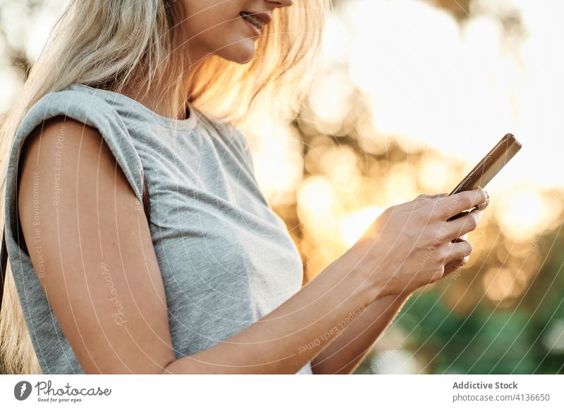 Anonyme Frau mit Smartphone im Park stehend benutzend genießen Browsen jung trendy Lifestyle Apparatur Gerät Kälte positiv soziale Netzwerke Lächeln online