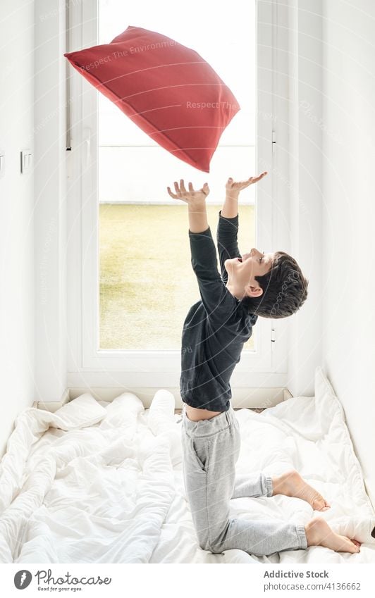 Fröhlicher Junge spielt mit Kissen auf dem Bett spielen Kopfkissen werfen Decke Freude Kind Spaß haben Wochenende spielerisch heimwärts gemütlich weich Stock