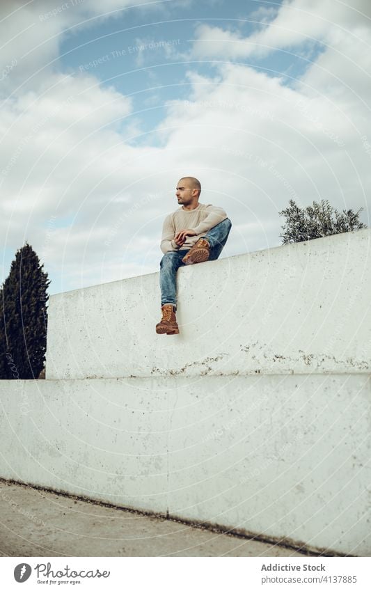 Hipster Mann sitzt auf Betonwand besinnlich Stil trendy modern Streetstyle nachdenklich nachdenken Zaun Wand jung unrasiert Typ Jeanshose sitzen lässig Himmel