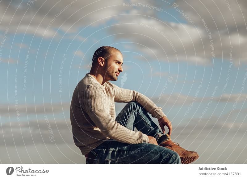 Hipster Mann sitzt gegen bewölkten Himmel besinnlich Stil trendy modern Streetstyle nachdenklich nachdenken Wolken sonnig jung unrasiert Typ Jeanshose Stiefel