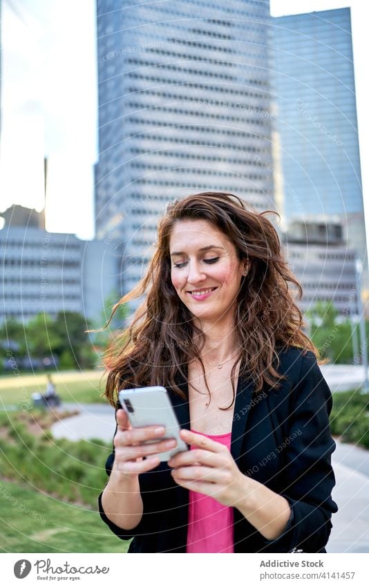 Fröhliche Geschäftsfrau in Jacke, die auf der Straße ihr Smartphone durchsucht Unternehmer benutzend Großstadt stilvoll Browsen Lächeln urban Frau Mobile