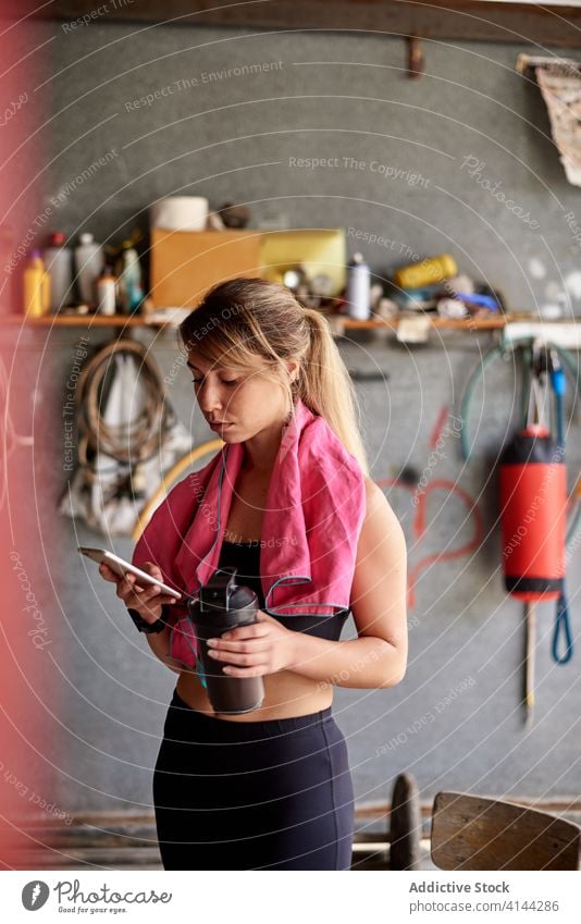 Konzentrierte Sportlerin mit Smartphone im Fitnessstudio in der Garage benutzend Fokus Konzentration Flasche Pause passen Apparatur Browsen Funktelefon Hydrat