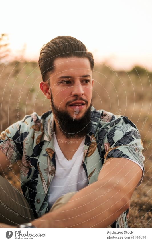 Nachdenklicher bärtiger Mann sitzt bei Sonnenuntergang im Gras auf einem Feld nachdenken Hipster Mund geöffnet Harmonie idyllisch Landschaft Himmel Wochenende