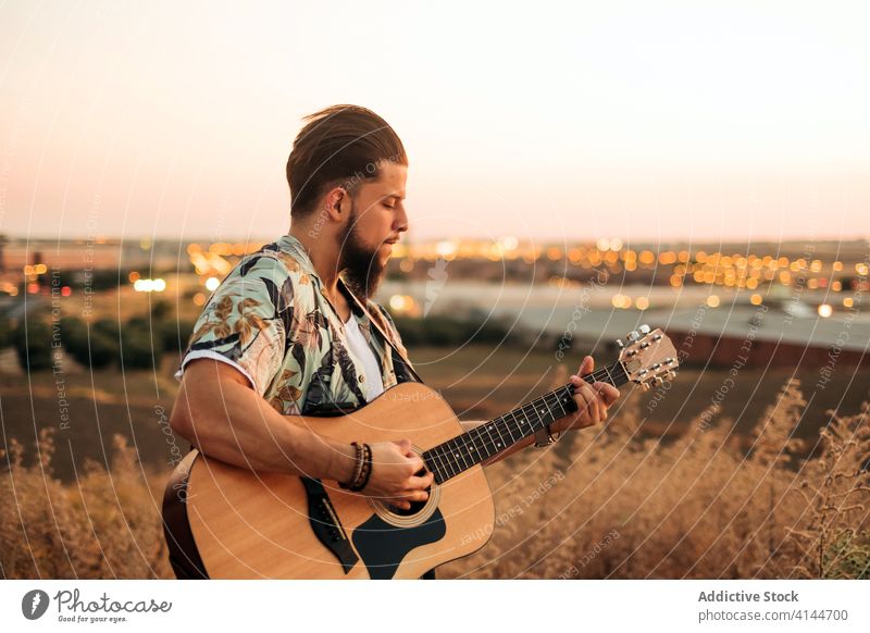 Männlicher Musiker spielt Gitarre auf dem Land bei Sonnenuntergang spielen Hipster Gras Himmel Feld Harmonie Mann Musical Instrument Gitarrenspieler künstlich