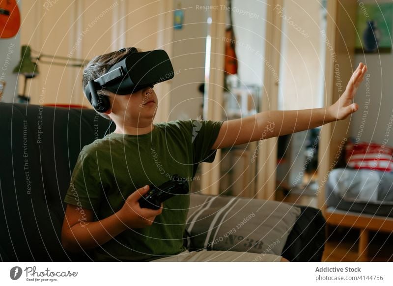 Kind in VR-Headset spielt auf der Couch Junge Schutzbrille spielen Gamepad virtuell Realität benutzend Sofa unterhalten modern heimwärts Gerät wenig bezaubernd